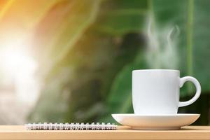 White coffee cup with stream and notebook on wooden table garden in background. photo