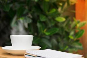 White coffee cup with stream and notebook on wooden table garden in background. photo