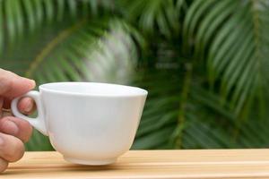 Man hand holding a white coffee mug with stream on a wooden table and palm tree in background.-2 photo
