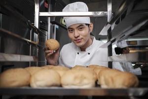 Young Asian male chef in white cook uniform and hat showing tray of fresh tasty bread with a smile, looking at camera, happy with his baked food products, professional job at stainless steel kitchen. photo