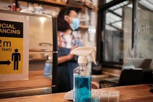 Alcohol sanitizer for healthy protection in a cafe, Asian male barista in face mask use laptop, waiting for coffee order in COVID19 pandemic quarantine, the economic impact for small business startup. photo