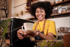 African American female barista in looks at camera, offers cup of coffee to customer with cheerful smile, happy service works in casual restaurant cafe, young small business startup entrepreneur. photo