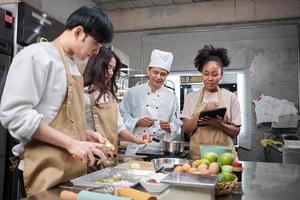 Hobby cuisine course, senior male chef in cook uniform teaches young cooking class students to peel and chop apples, ingredients for pastry foods, fruit pies in restaurant stainless steel kitchen. photo