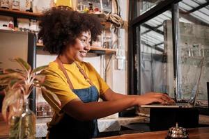 African American female cafe barista in casual apron happy with online customer order in laptop, cheerful works with smile at counter bar in coffee shop, small business startup entrepreneur lifestyle. photo