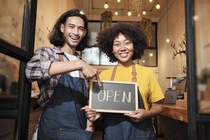 dos jóvenes socios baristas de inicio con delantales se paran en la puerta de un café informal, cartas a bordo y muestran un cartel abierto, sonrisas felices y alegres con trabajos de servicio de cafetería y nuevos empresarios. foto