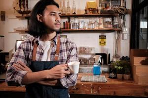 Long hair handsome Asian male startup barista with an apron stands at a casual cafe, arms crossed, looking outside with a coffee cup, alcohol sanitizer on the counter bar, coffee shop service jobs. photo