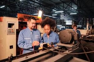 Asian male and female African American engineers in safety uniform work by inspecting machines' voltage current, checking, and maintaining at manufacture factory, electric system service occupations. photo