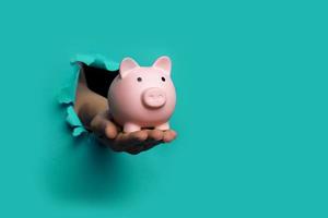 hand holds a piggy bank blue background out of a torn hole in a paper background photo
