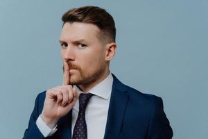Serious bearded prosperous male office worker dressed in formal clothes makes hush gesture asks to keep quiet demands silence with strict expression isolated over blue background. Taboo shh shushing photo