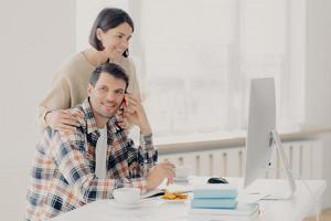 Happy husband and wife do paperwork together, pay taxes online look at screen computer, pose at desktop with books and papers, drink coffee and have lunch, make telephone call via smartphone photo