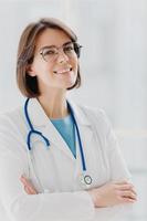 Close up portrait of short haired female general practitioner stands with smile and arms crossed, uses stethoscope, enjoys work, poses against white background. People, mediccare and treatment concept photo