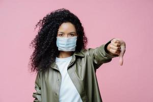 Image of serious curly haired young woman wears medical mask, keeps thumb down, shows dislike gesture, avoids spreading of corona virus, cares about health. Dangerous virus. Medical concept. photo