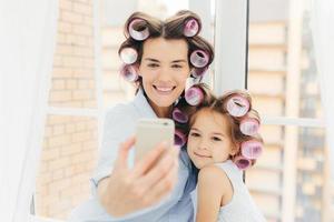 Indoor shot of pleasant looking mum with positve expression and her daughter do curly hairstyles, pose for selfie in modern cell phone, prepare for carnival or party. Women and beauty concept photo