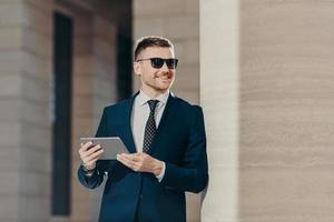 Portrait of handsome young male employer holds digital tablet, connected to wireless internet, wears sunglasses and formal suit, has free time after hard working day. People and career concept