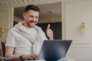 Happy young man freelancer in earphones showing thumb up while talking by video chat with colleague at home office photo