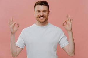 Young satisfied bearded man makes okay gesture with both hands expresses agreement approves something praises nice work says well done dressed in casual white t shirt isolated on pink studio wall photo