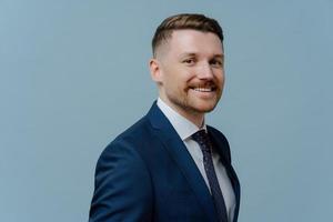 Sideways shot of smiling corporate worker dressed in formal outfit comes on business meeting poses against blue studio background copy space aside. Elegant businessman wears office clothing. photo