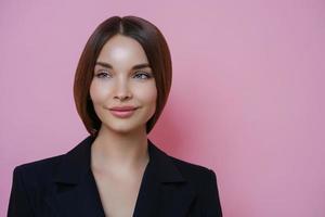 hermosa mujer de negocios mira a un lado con expresión soñadora, piensa en algo agradable durante el descanso después del trabajo, usa ropa negra, aislada en un fondo rosa, espacio vacío para su información foto