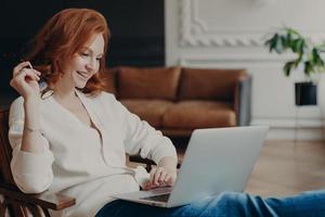 Skilled positive female student creats article for homework, sits in coworking space, has pleased expression, works on laptop computer, pose over home interior, prepares for writing course work photo