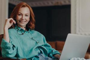 Indoor shot of pleasant looking redhead woman has good time at home, works on computer, wears casual shirt, holds eyeglasses, feels comfort, enjoys domestic atmosphere. Teacher teaches by video call photo