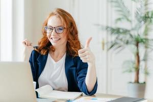 Positive redhead woman keeps thumb raised, demonstrates like gesture, satisfied with good work of colleague, updates software on modern gadget, searches data on website, poses in coworking space photo