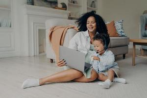 Happy small kid boy using headset while talking online on laptop, sitting on floor with mom photo