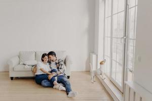 Satisfied husband and wife have romantic relationship, sit on floor near white couch in big room, wear jeans, tshirts and socks, spend leisure time in domestic atmosphere, dog poses near balcony photo