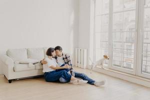 Horizontal view of happy affectionate family couple dressed in casual wear, embrace and express love to each other, pose on floor near sofa in modern apartment, their pet looks through big window photo
