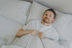 Lonely upset young bearded man lying in bed with open eyes photo
