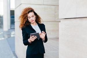 Elegant lady with curly hair, wearing black suit, typing messages or making business report while using tablet computer. Female entrepreneur working on new business project. Business and career photo