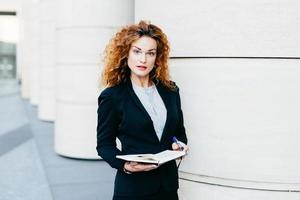Horizontal portrait of serious pretty businesswoman with curly hair, thin eyebrows and curly hair, wearing black suit and white blouse, holding notebook with pen in hands, looking directly into camera photo