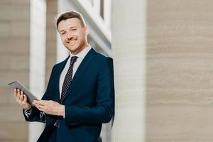 Portrait of stylish male entrepreneur has cheerful expression, holds digital tablet, reads financial news on web page, uses free internet connection, wears formal suit and tie. Career concept photo