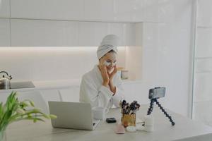 Female beauty blogger in bathrobe and towel on head recording video on smartphone for her vlog photo