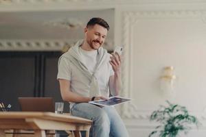 Happy male freelancer looking at phone and smiling while sitting on work desk photo