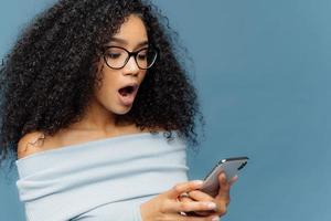 Shocked African American woman gots message from ex, gasps from surprise and excitement, reads stunning news on mobile phone, has curly bushy hair, wears jumper, isolated over blue studio wall photo