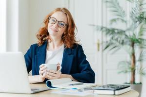 Pleased female teacher makes research work on laptop computer, writes down information in notepad, enjoys drinking coffee, has happy smile on face, sits in spacious room at desktop plant in background photo