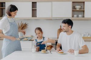 la familia desayuna en la cocina por la mañana. la niña feliz pone chocolate derretido en un sabroso panqueque frito, posa en la mesa con el padre, el perro, la madre se para cerca, usa delantal, sostiene la sartén, cocina ocupada foto