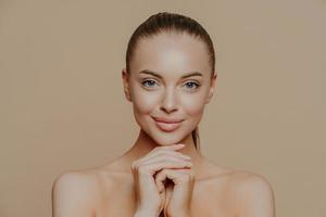Young pretty woman with beauty skin and combed hair, keeps hands under chin, has healthy complexion after face purification, soft well cared body, isolated over studio wall. Facial treatment photo