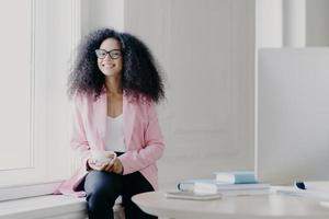 la empresaria feliz tiene el pelo afro tupido, usa una chaqueta rosada y pantalones negros, sostiene una taza de bebida, posa en el alféizar de la ventana en un espacioso gabinete con mesa, computadora moderna, disfruta de un café aromático foto