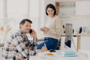 Horizontal shot of cheerful man has telephone conversation, discusses financial contract agreement, dressed in checkered shirt, writes down information, happy woman holds mug of drink, leans at table photo