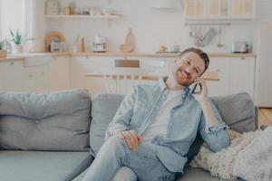 Smiling guy sits on couch at home having fun while talking on modern cellphone gadget photo