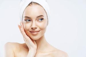 hermosa modelo femenina con maquillaje, toca la mejilla, mira a un lado, tiene la piel limpia y fresca después de bañarse, usa una toalla suave en la cabeza, aislada sobre fondo blanco, copia espacio. cuidado facial, concepto de higiene foto