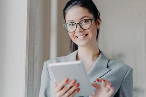 Indoor shot of happy brunette entrepreneur reads electronic book on modern digital tablet, reads financial news in internet during work break, wears optical glasses, grey costume, poses indoor photo