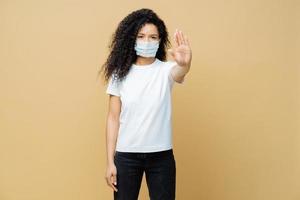 Say no to spreading disease. Serious looking Afro American woman makes stop gesture, wears medical mask, prevents virus Covid-19, stands indoor, isolated. Stop infectious disease immediately photo