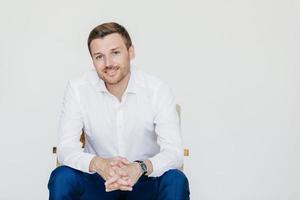 Studio shot of elegant cheerful male entrepreneur in formal clothes, sits on chair, poses at camera, isolated over white background. Glad man smiles happily, rejoices his success. Office worker photo