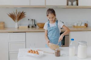 Cute small girl wears striped apron, whisks ingredients in bowl, prepares dough, teaches to cook, stands at white table with eggs, milk, flour against kitchen background. Childern and cooking concept photo