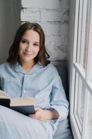 Photo of beautiful woman dressed in casual clothes, wears pyjamas, sits on window sill, reads romantic story in book, enjoys coziness and comfort. Young female with textbook at home. Leisure concept