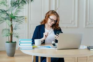 Smiling red haired female focused in modern cellular, happy to recieve text message, poses at workplace, drinks coffee, works on financial project gets ready for video conference with business partner photo