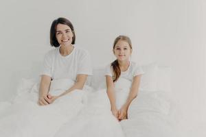 Morning time and awakening concept. Happy smiling mother and daughter sit in comfortable bed, dressed in white t shirts, awake with positive mood, glad to start new day, spend free time in bedroom. photo