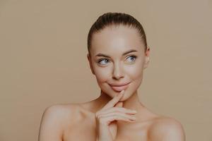 Pensive attractive young European woman with white healthy skin, looks aside, has naked body, undergoes beauty treatments, poses against beige background, has combed dark hair, bare shoulders photo